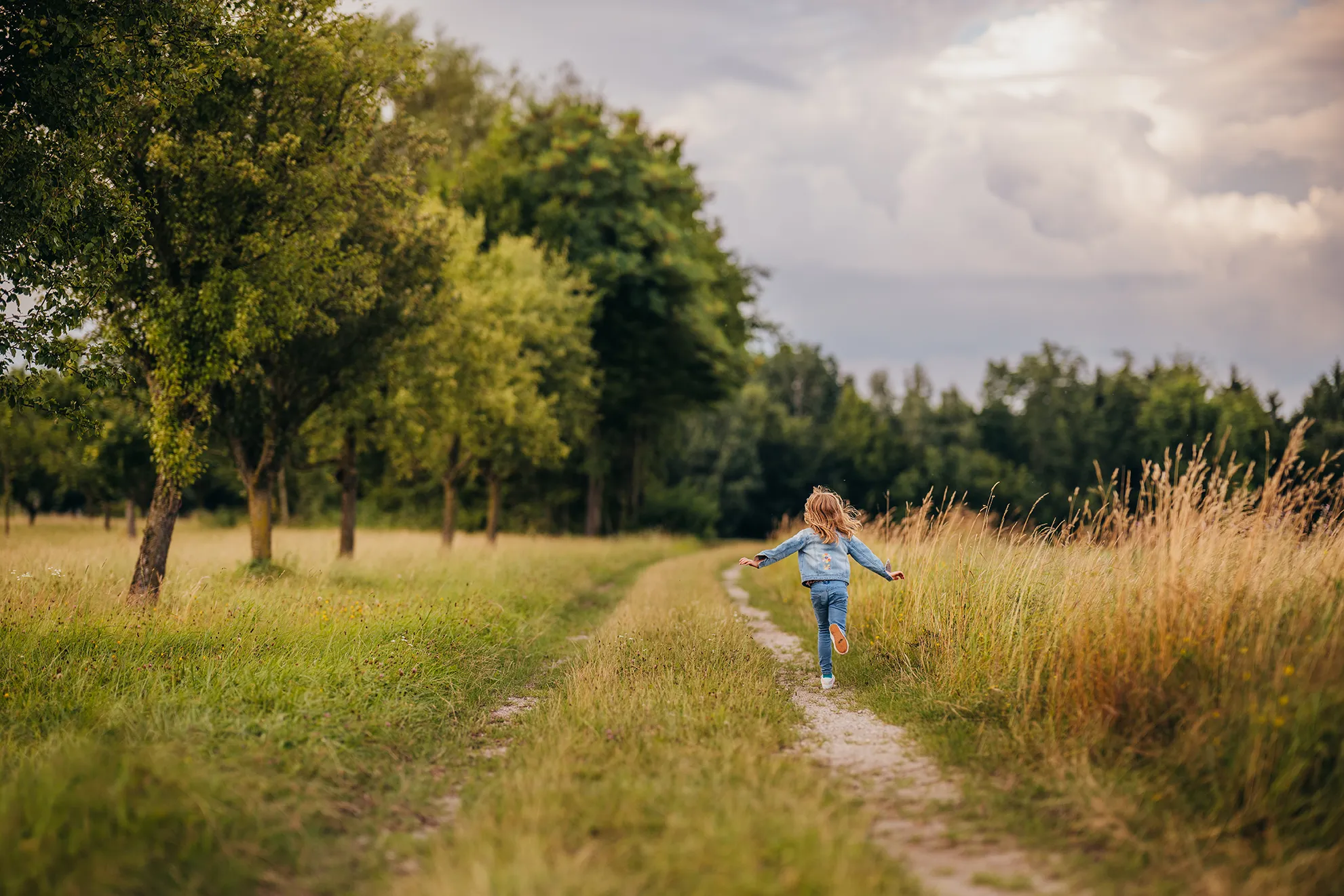 Ein Mädchen läuft zwischen Obstbäumen und Gras über einen Feldweg, der Himmel zeigt Gewitterstimmung